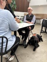 Image of 2 people sitting at a cafe table. A harnessed guide dog lays on the ground next to them.
